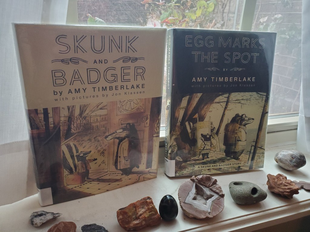 photo of the books "Skunk and Badger" and "Egg Marks the Spot" on a windowsill. There are various rocks scattered in front of them.