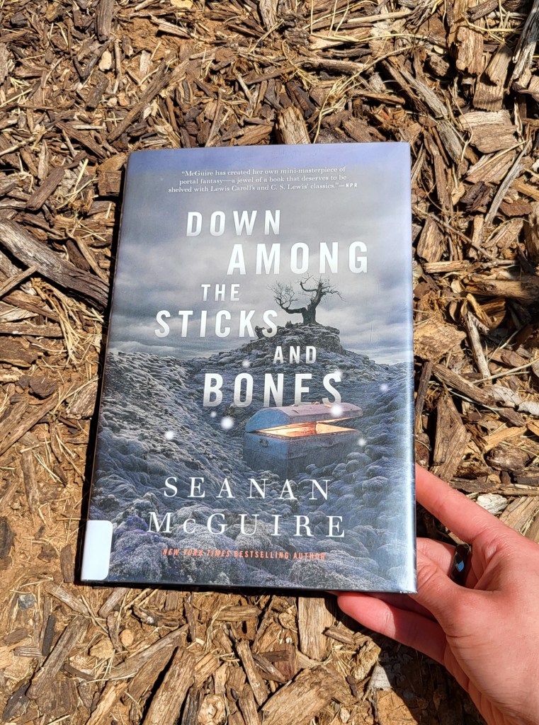 photo of Down Among the Sticks and Bones being held over a garden filled with mulch and twigs. The book cover depicts a desolate landscape, a gnarled tree in the distance, and a trunk half-open, glowing with otherworldly light, in the foreground.