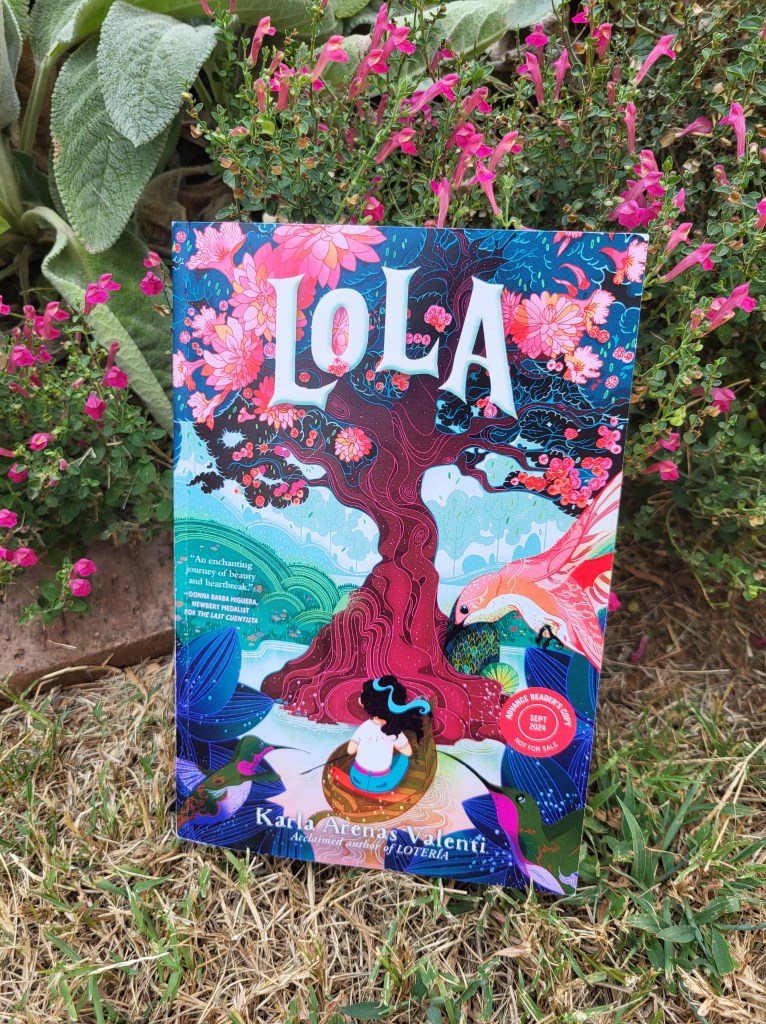 photo of Lola: a tree with pink blossoms dominates the cover, with a boat carrying a young girl sits at the base of the tree. The book itself rests against a flowering plant in a garden.