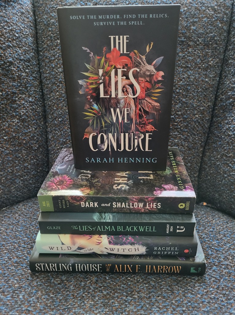 photo of The Lies We Conjure: the cover depicts various objects (plants, a human heart, a vial of blue liquid, a rabbit, etc.) on a black background. The book itself on a stack of other books (Dark and Shallow Lies, The Lies of Alma Blackwell, Wild is the Witch, and Starling House).
