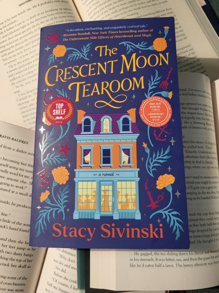 photo of the cover of The Crescent Moon Tearoom, which depicts a building with glowing windows with the silhouettes of people. Around the building is a border of leaves and flowers. The book itself rests on the open pages of other fantasy novels.