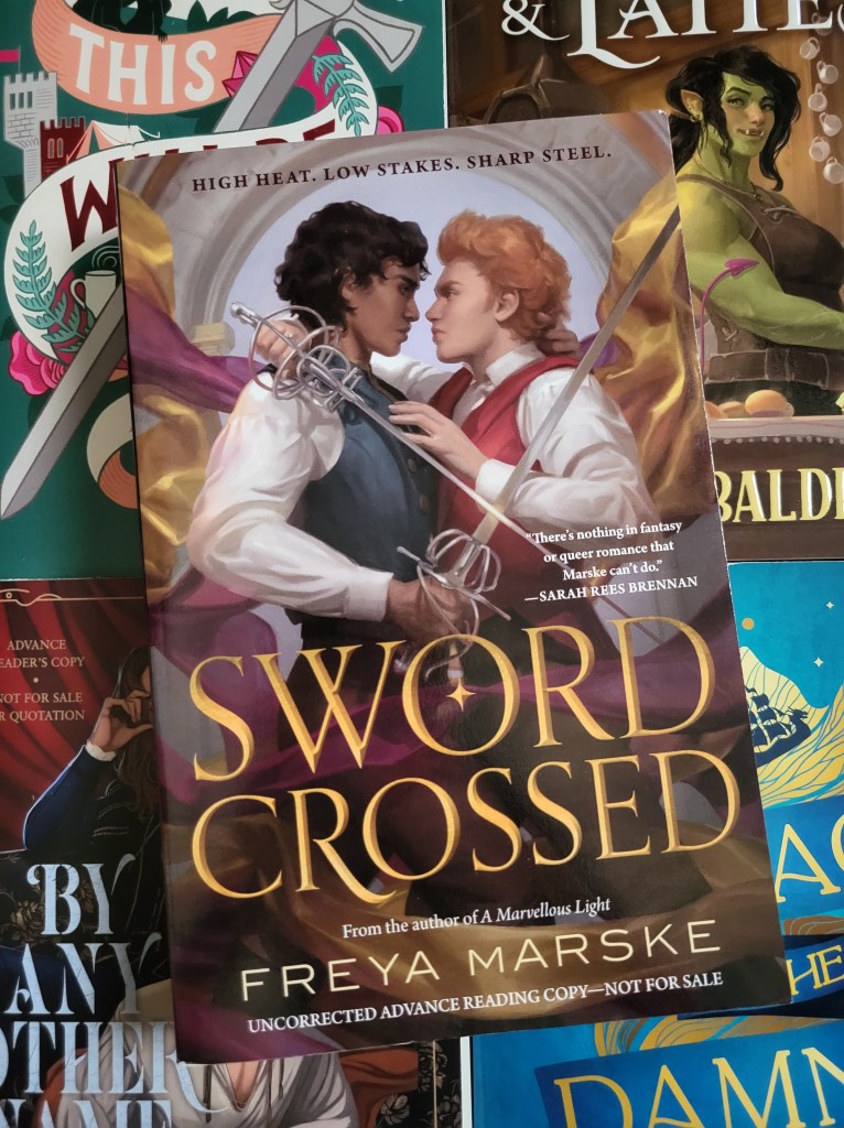 photo of Swordcrossed: two young men in waistcoats and white shirts have their arms around each other, swords in their hands. Rippling swathes of gold and purple fabric are behind and around them. The book itself sits on other fantasy romance books (This Will Be Fun, Legends & Lattes, By Any Other Name, and Voyage of the Damned)