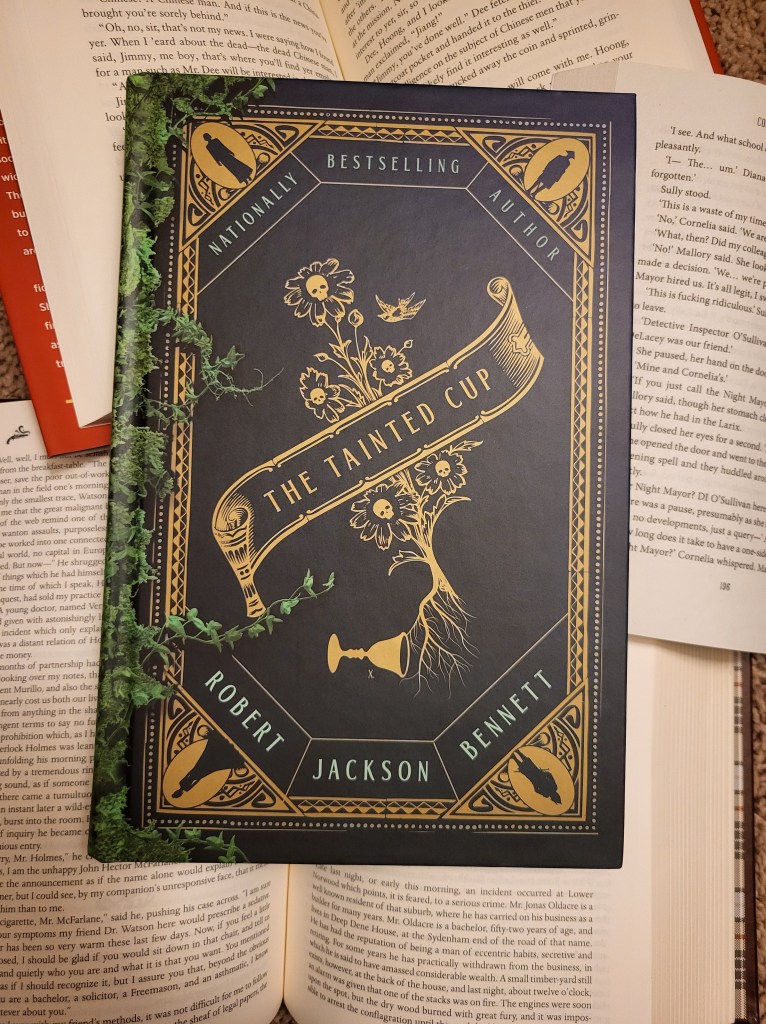 photof the Tainted Cup: a golden border outlines the cover, with silhouettes of people in each corner. In the center of the cover is the title written on a ribbon, with curling flowers and skulls behind it. The flowers bloom from a cup, which has been tipped over. On the spine of the book, spreading onto the front like grasping tendrils, there are green vines.