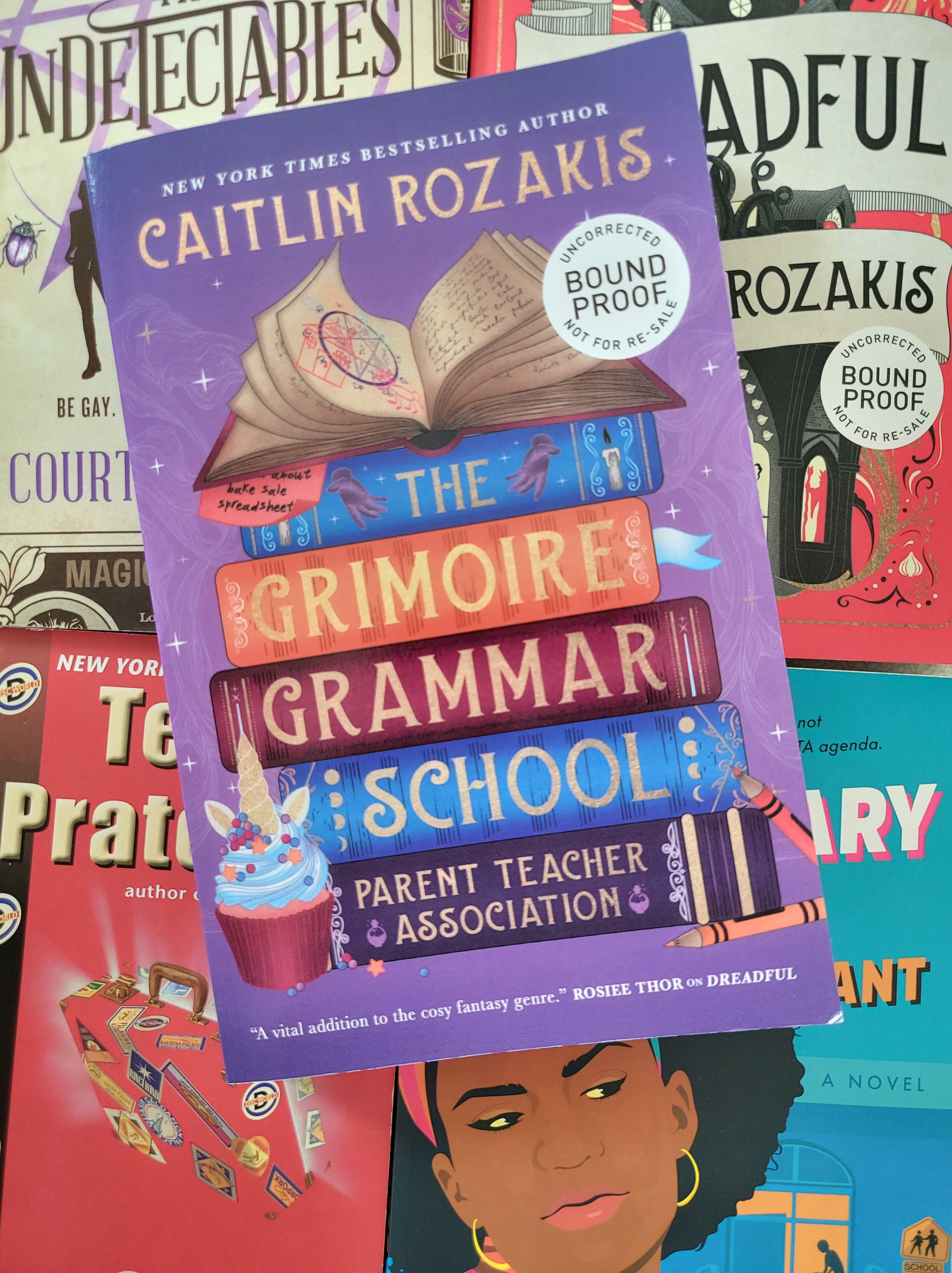 photo of Grimoire Grammar School: the cover is purple, depicting a stack of spell books. There are crayons and a unicorn cupcake next to the books. The novel itself is on top of other similar books: The Undetectables, Dreadful, The Color of Magic, and It's Elementary.