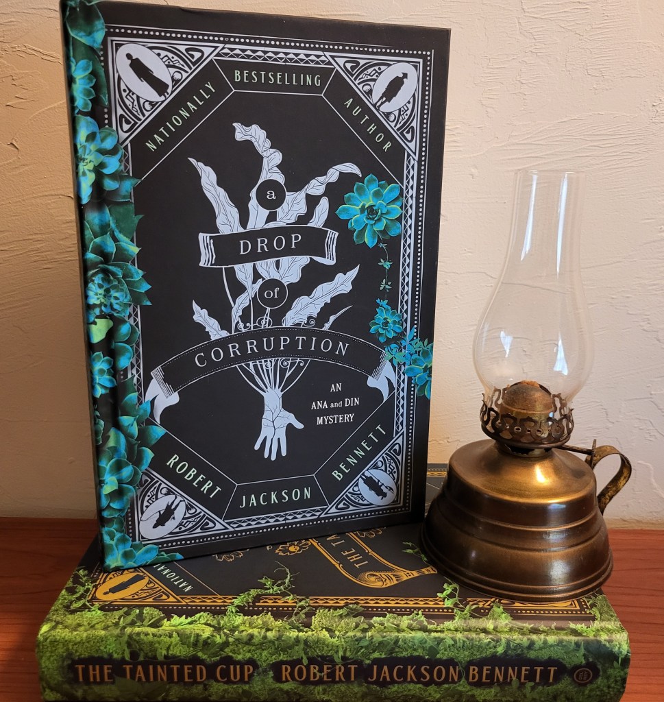 photo of A Drop of Corruption: a black background with a silver border, depicting the silhouettes of four people. In the center of the cover is a bundle of silver leaves, over which the title is written. At the edges of the cover are green succulent plants. The book itself sits on top of its predecessor The Tainted Cup.