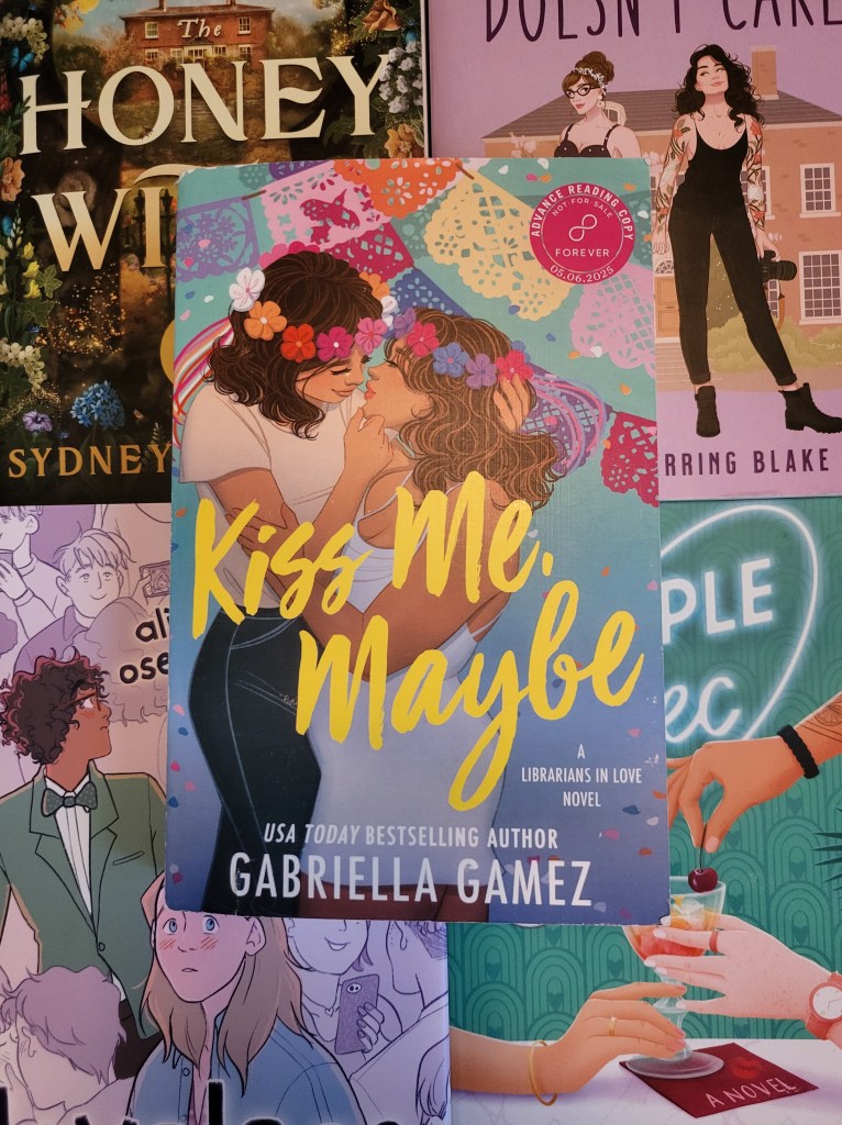 photo of Kiss Me Maybe: a blue background with colorful papel picado hanging around. Two women embrace in the foreground. One wears a flower crown in the colors of the lesbian pride flag, the other the bisexual pride flag. The title appears superimposed over them in yellow letters.