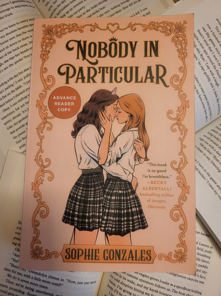 photo of Nobody in Particular: an ornate border in gold, over a pinkish background. Two teens, one brunette and one blonde, embrace in the center of the cover. They're wearing white button-ups and plaid skirts, and the brunette has a delicate tiara on her head.