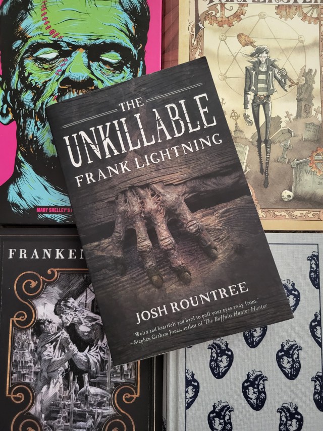 photo of Unkillable Frank Lightning: a gnarled, rotting hand gripping parts of what appears to be a wooden fence. The title and author's name are in white above and below this image. The book itself rests at an angle on top of four different editions of Frankenstein.