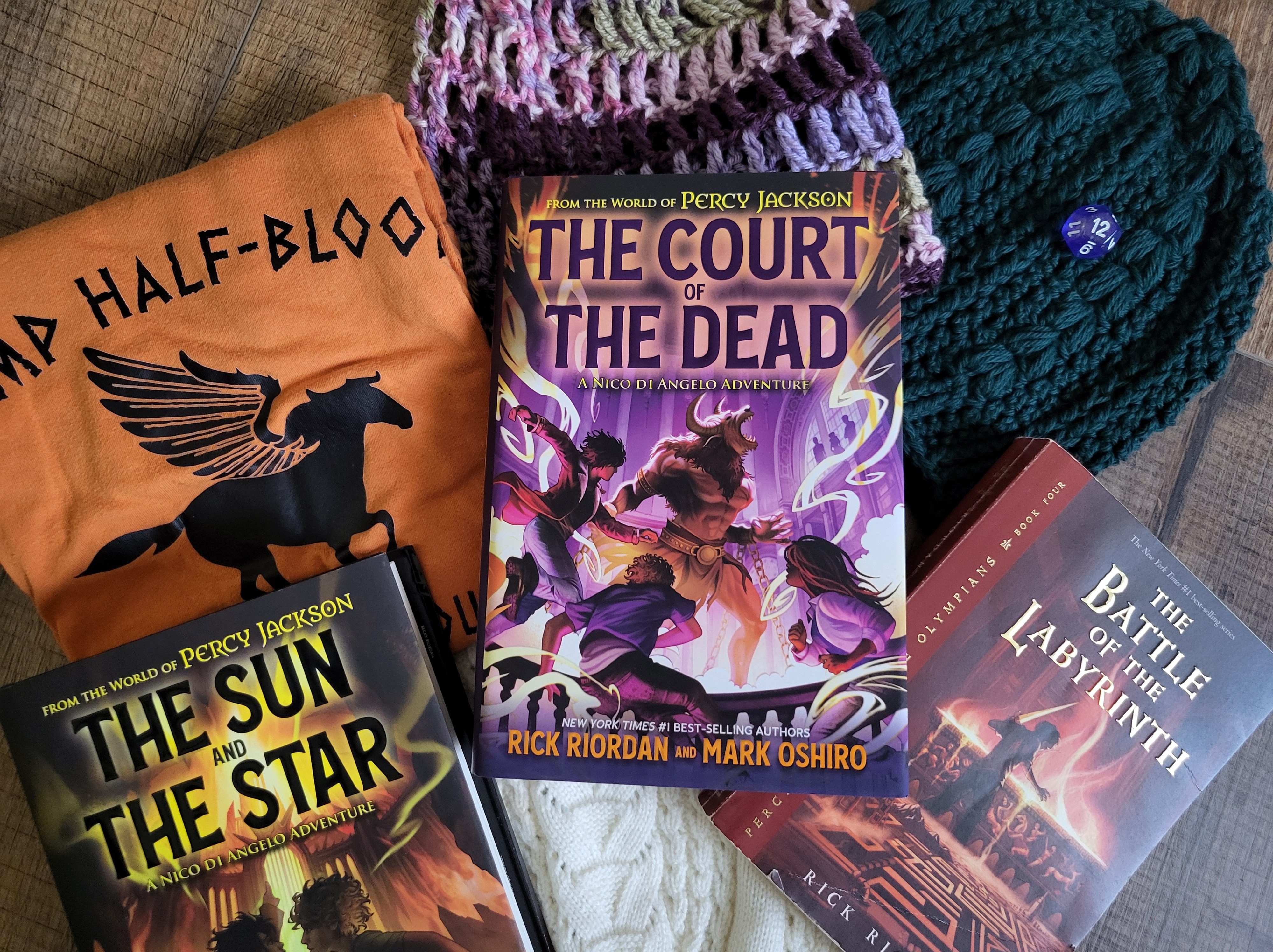 photo of The Court of the Dead: the cover depicts three teens surrounding the Minotaur, a half-man, half-bull creature. Purple lights swirls behind them all, and the title is in dark purple text outlined in orange. The book itself sits on several knitted hats and a Camp Half-Blood t-shirt. The books The Sun and the Star and Battle of the Labyrinth are also nearby, as is a twelve-sided dice. All of these objects make sense in the context of the book, I promise.