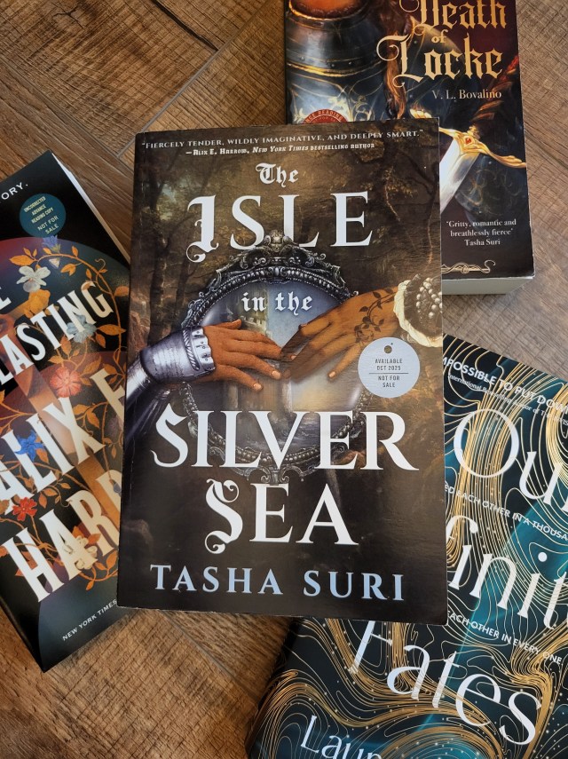 photo of Isle in the Silver Sea: the background is dark brown with trees visible. There's a silver framed mirror, and over that are two hands reaching for each other. One has leaves tattooed across the skin and a lace sleeve, the other wears silver plate armor. The title is in white. The book itself sits on similar titles: The Everlasting, The Second Death of Locke, and Our Infinite Fates.