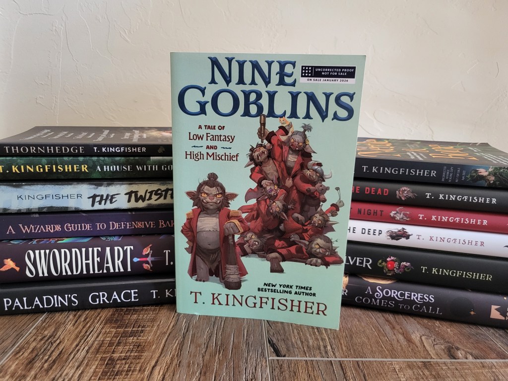 Photo of Nine Goblins: a pale green cover, on which there is a pile of eight goblins wearing red jackets. They brandish weapons, cuddle a teddy bear, and clutch a kitten. A ninth goblin stands to the left, clutching a wooden pace. The book itself stands in front a stack of other Kingfisher books.