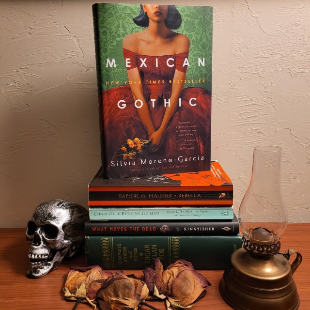 photo of Mexican Gothic: the book has a green brocade wallpaper background, with a woman in a red dress clutching flowers in front of it. The book itself sits on a stack of others (Rebecca, The Yellow Wallpaper, What Moves the Dead, and a Poe collection). There are dried roses, a fake skull, and a lantern around the books.
