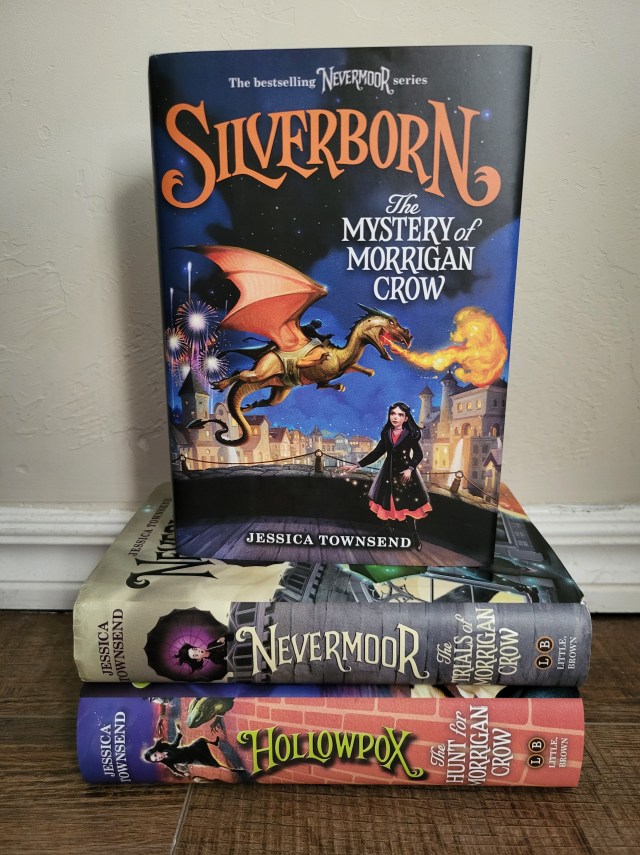 photo of Silverborn: the cover depicts an orange-winged dragon flying over a city. A young girl with brown hair looks up at it in the foreground. The book itself sits on two others in the series.