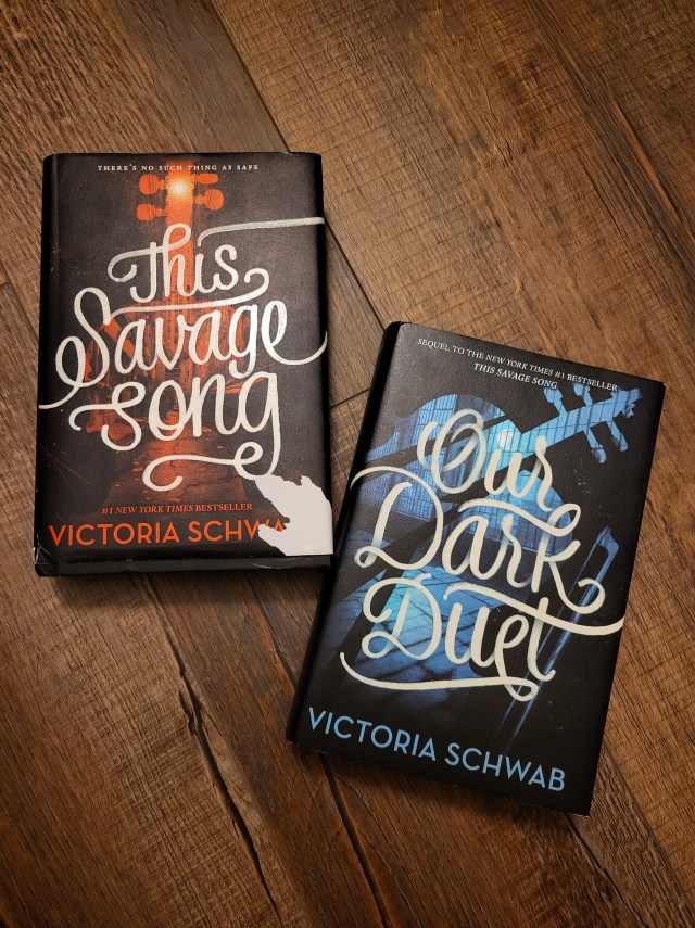 photo of This Savage Song and Our Dark Duet: both books have a black background, the former with a red silhouette of a violin and the latter with a blue one. The titles are in curling white letters over these violins.
