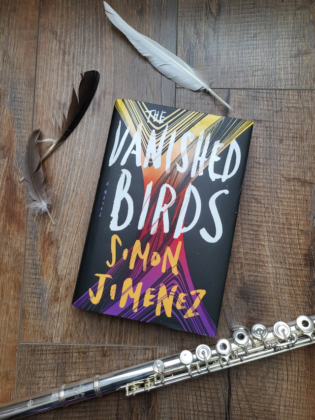 photo of The Vanished Birds: a black background with a spiral of yellow, orange, red, and purple. The cover (in white) and the author's name (in orange) are atop the spiral. The book itself sits on a brown background, with several features and flute surrounding it.