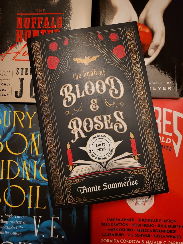photo of The Book of Blood and Roses: a black background with gold floral framing, and red roses. A bat silhouette is at the top, and an open book with candles is at the bottom. The title is in white in the center of the cover. The book itself is sitting on other vampire books (The Buffalo Hunter Hunter, Twilight, Bury Our Bones in the Midnight Soil, and Vampires Never Get Old)