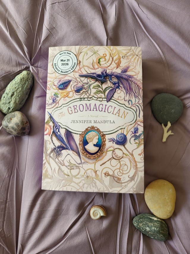 photo of The Geomagician: the book has a cream colored background, with various fossils pictures. There is also a cameo brooch of a woman beneath the title, which is in purple letters in the center of the cover. The book itself rests on pale purple fabric, with stones and shells around it.