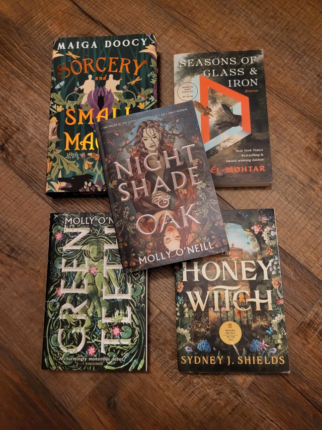 photo of Nightshade and Oak: plants surround two women, one with brown hair and the other read. The latter is upside down on the cover, giving the image symmetry. The title is in silver letters over the image. The book itself sits on other fantasy/romance books (Sorcery & Small Magics; Seasons of Glass and Iron; Greenteeth; and The Honey Witch).