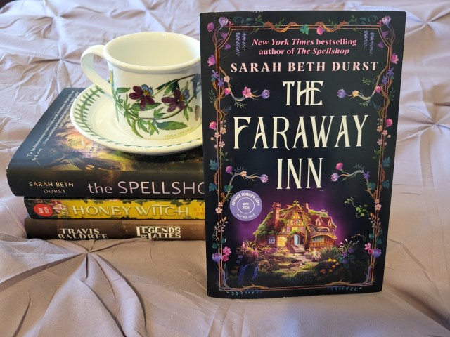 photo of The Faraway Inn: The title is on the top half of the cover, with a small ivy covered stone building on the bottom. Winding vines and flowers make up the border. The book itself leans against a stack of other novels (The Spellshop, The Honey Witch, and Legends & Lattes). A teacup with purple flowers is atop this stack.